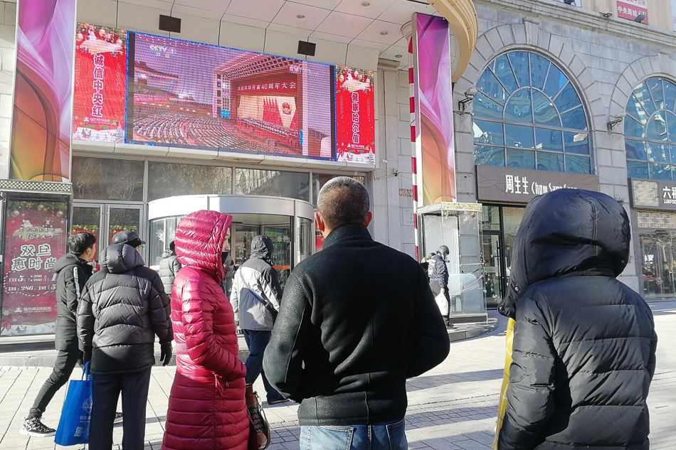 03-2018年12月18日，哈尔滨市道里区中央大街上，一家商场的大屏幕上播放庆祝改革开放40周年大会的电视直播，吸引了许多市民和游客观看。